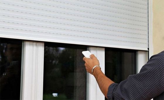 Persona que opera un control remoto blanco, probablemente para subir o bajar una persiana exterior blanca enrollable en una ventana.
