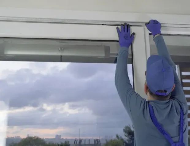 Persona con uniforme de trabajo azul y guantes instalando un marco de ventana en un interior, con un cielo nublado visible en el exterior.