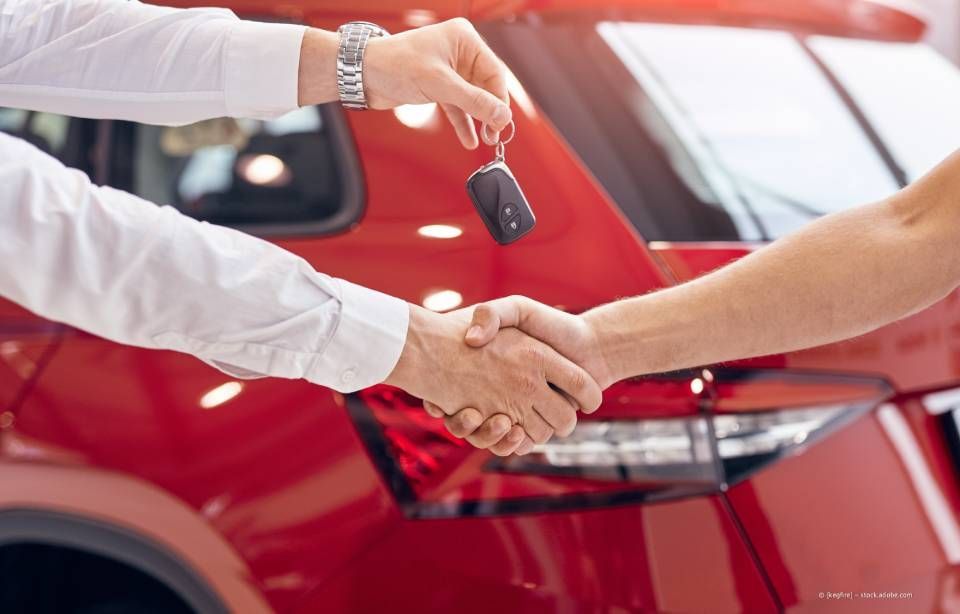Vor einem roten Auto zittern Hände, eine hält einen Schlüssel.