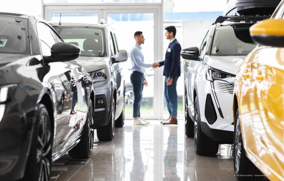 Zwei Männer schütteln sich in einem Autohaus, umgeben von Neuwagen, die Hände.