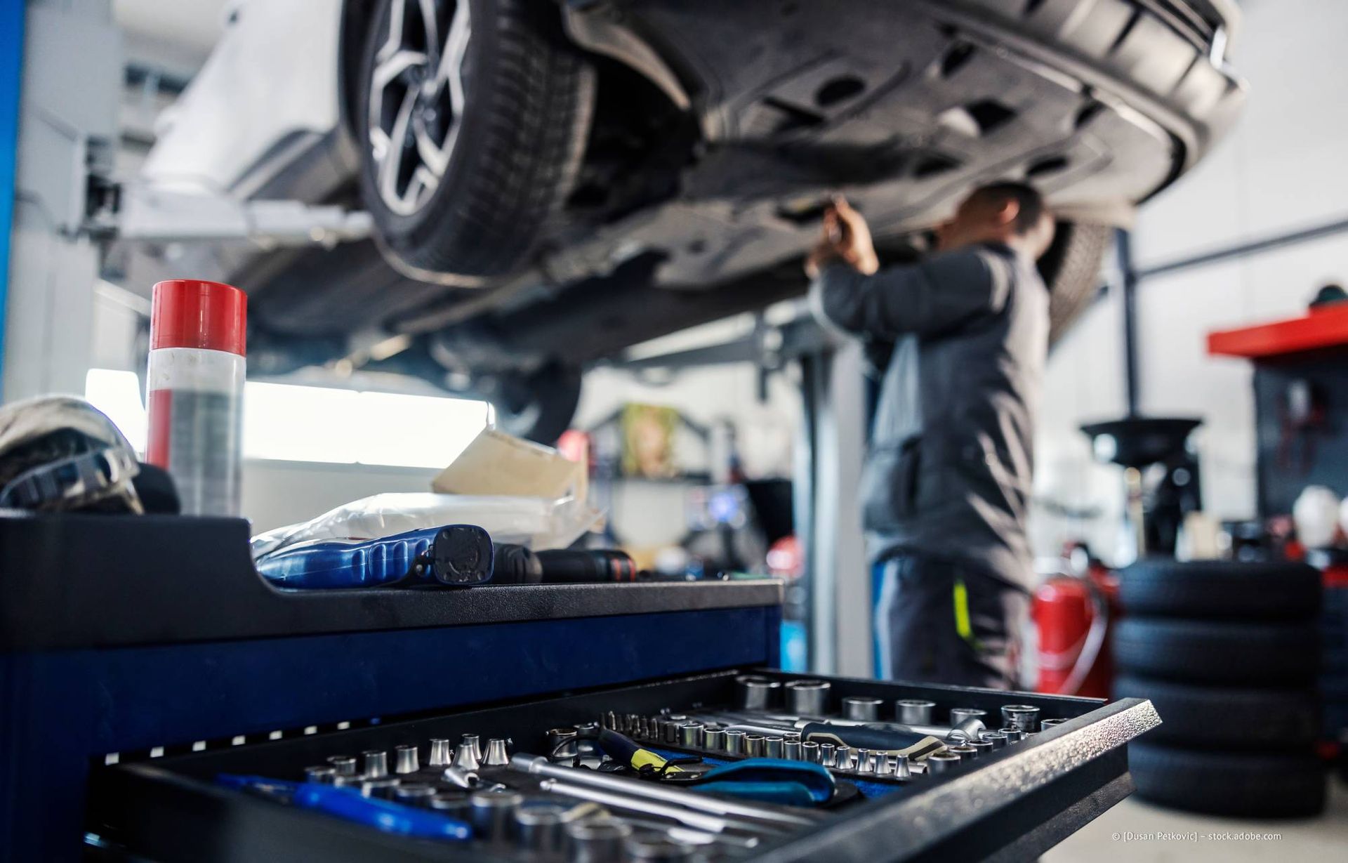 Ein Mechaniker arbeitet in einer Werkstatt an einem auf einer Hebebühne angehobenen Auto; Werkzeuge sind im Vordergrund zu sehen.