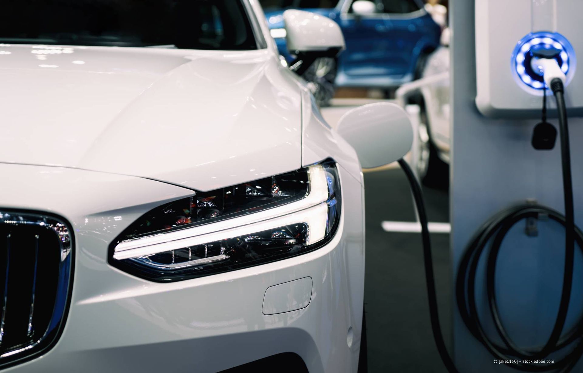 Ein weißes Elektroauto lädt an einer Ladestation. Schwarzes Kabel ist eingesteckt; blau-weißes Ladegerät ist sichtbar.