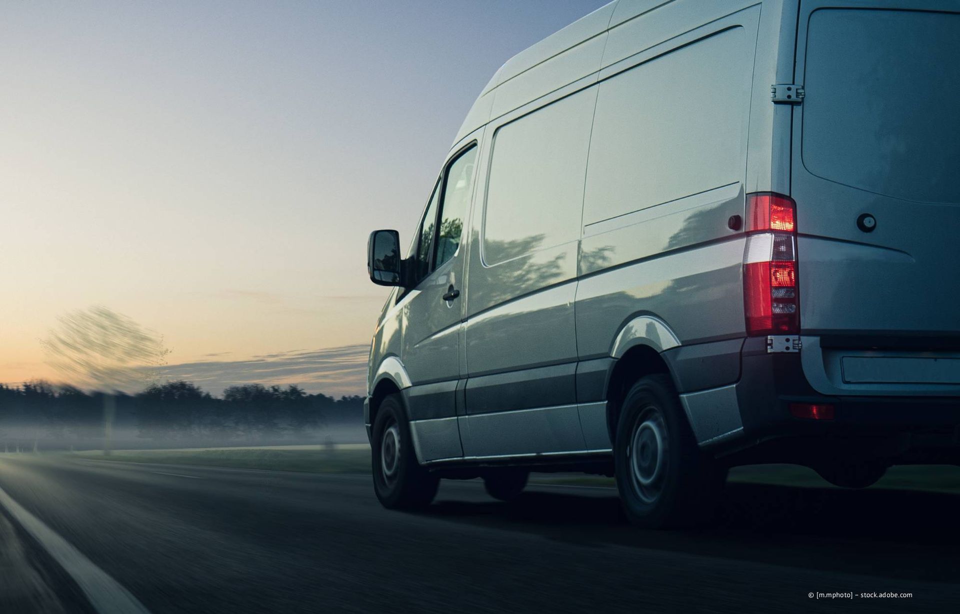 Ein weißer Lieferwagen fährt in der Abenddämmerung auf einer Straße; die Rücklichter sind an, im Hintergrund sind Bäume zu sehen.