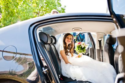 La mariée, assise dans une voiture décorée, sourit et tient un bouquet, vêtue d'une robe de mariée blanche.