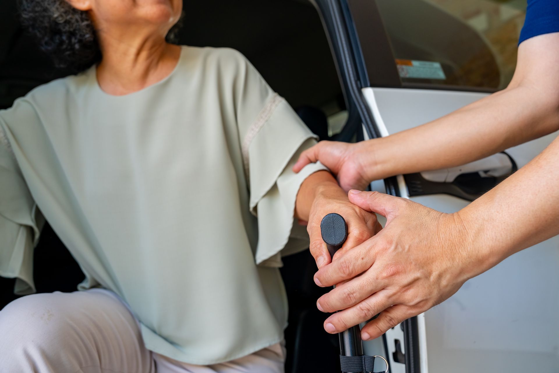 Une personne soignante aide une personne assise à sortir de la voiture en lui tenant le bras pour la soutenir.