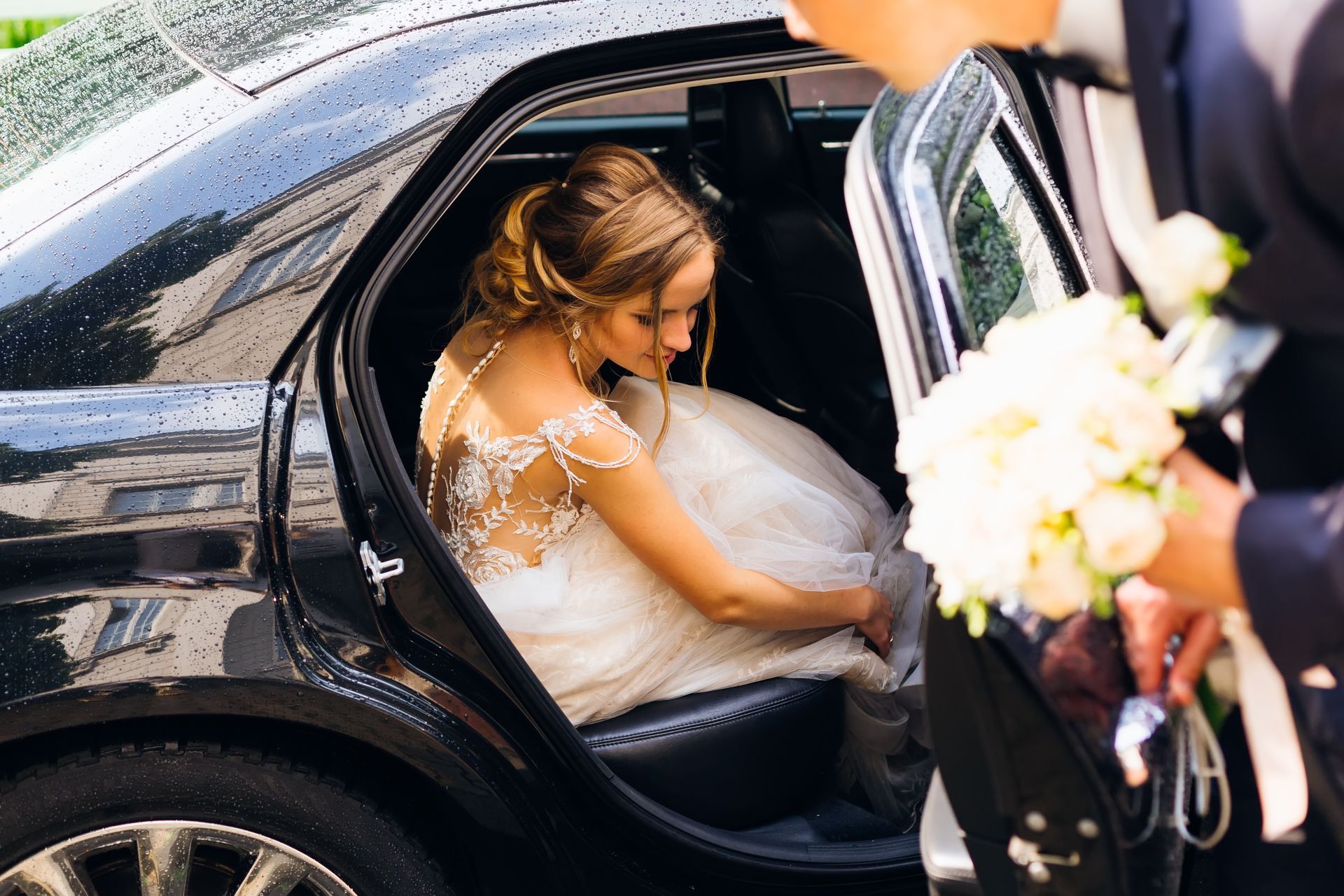 La mariée, assise sur le seuil d'une limousine noire, ajuste sa robe de mariée blanche tandis qu'une personne tient un bouquet à proximité.