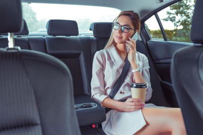 Une femme en robe blanche, assise sur la banquette arrière d'une voiture, tient un téléphone à l'oreille.