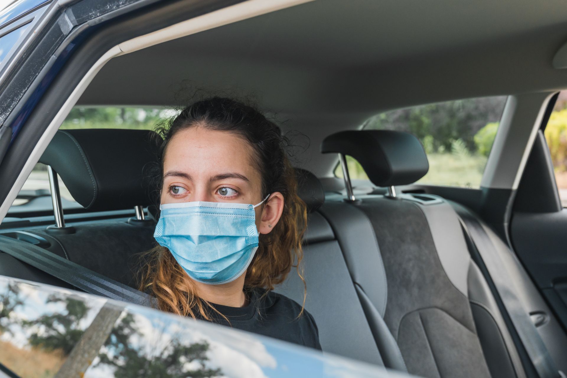 Une femme portant un masque bleu est assise sur la banquette arrière d'une voiture, regardant par la fenêtre.