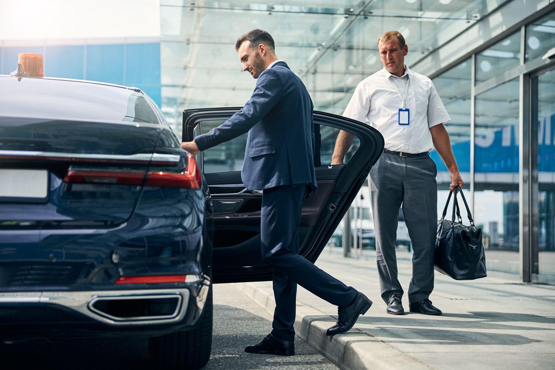 Deux hommes près d'une berline noire garée sur le trottoir d'un aéroport, l'un d'eux ouvrant le coffre et portant un sac.