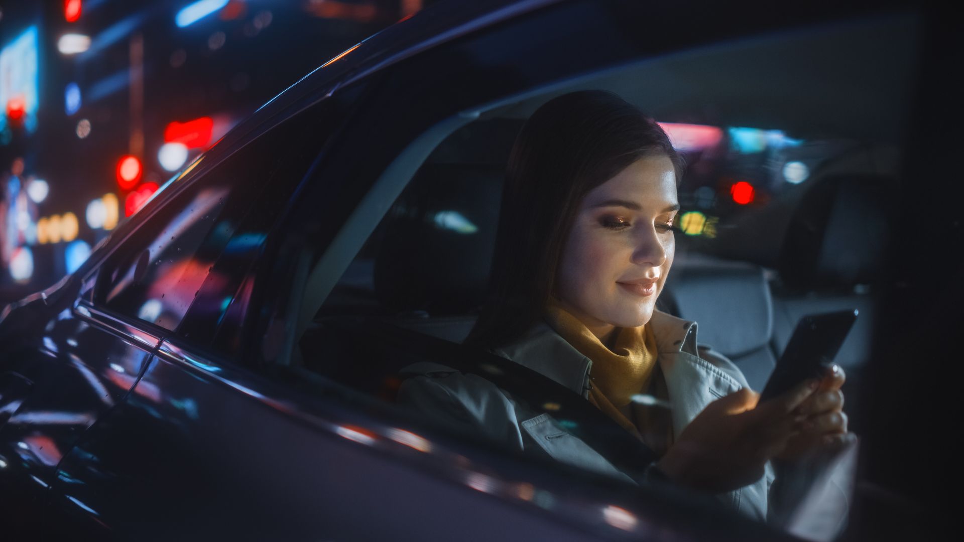 Une femme utilise son téléphone portable dans une voiture la nuit, les lumières de la ville se reflétant sur la vitre.
