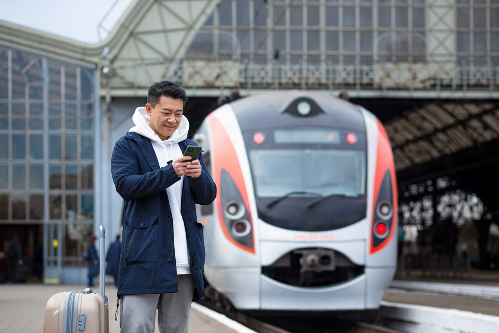 Un homme regarde son téléphone sur le quai d'une gare tandis qu'un train blanc et rouge approche derrière lui.