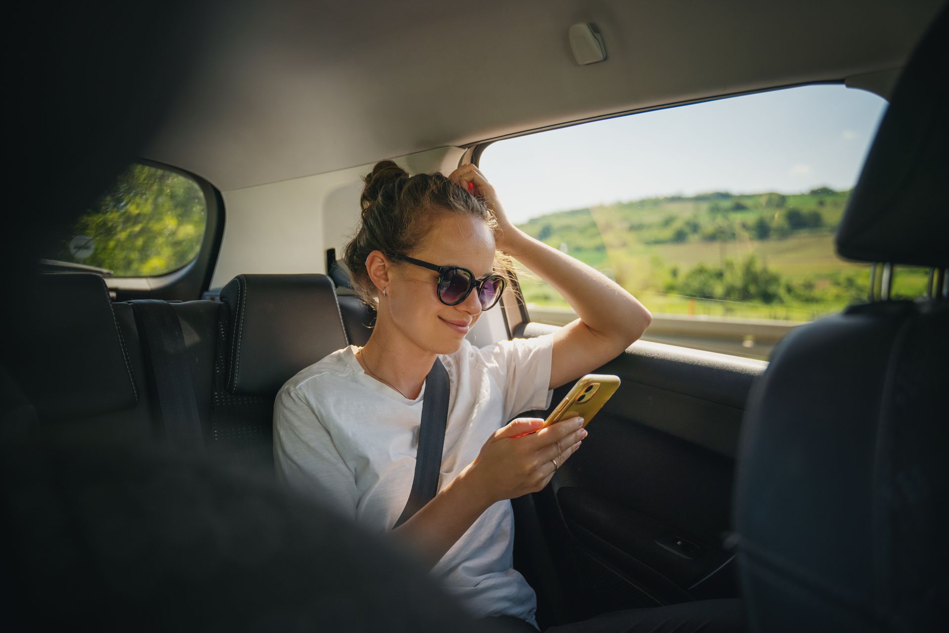 Une femme portant des lunettes de soleil utilise son téléphone sur la banquette arrière d'une voiture, avec un paysage ensoleillé à l'extérieur.