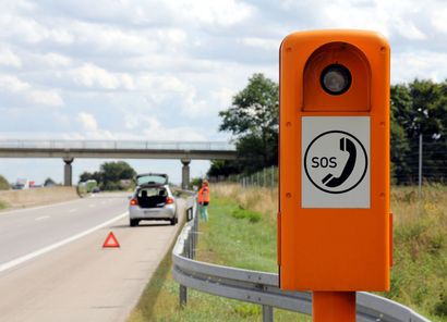 Téléphone d'urgence orange en bord de route, sur le bas-côté de l'autoroute, une voiture arrêtée à proximité et un pont en arrière-plan.