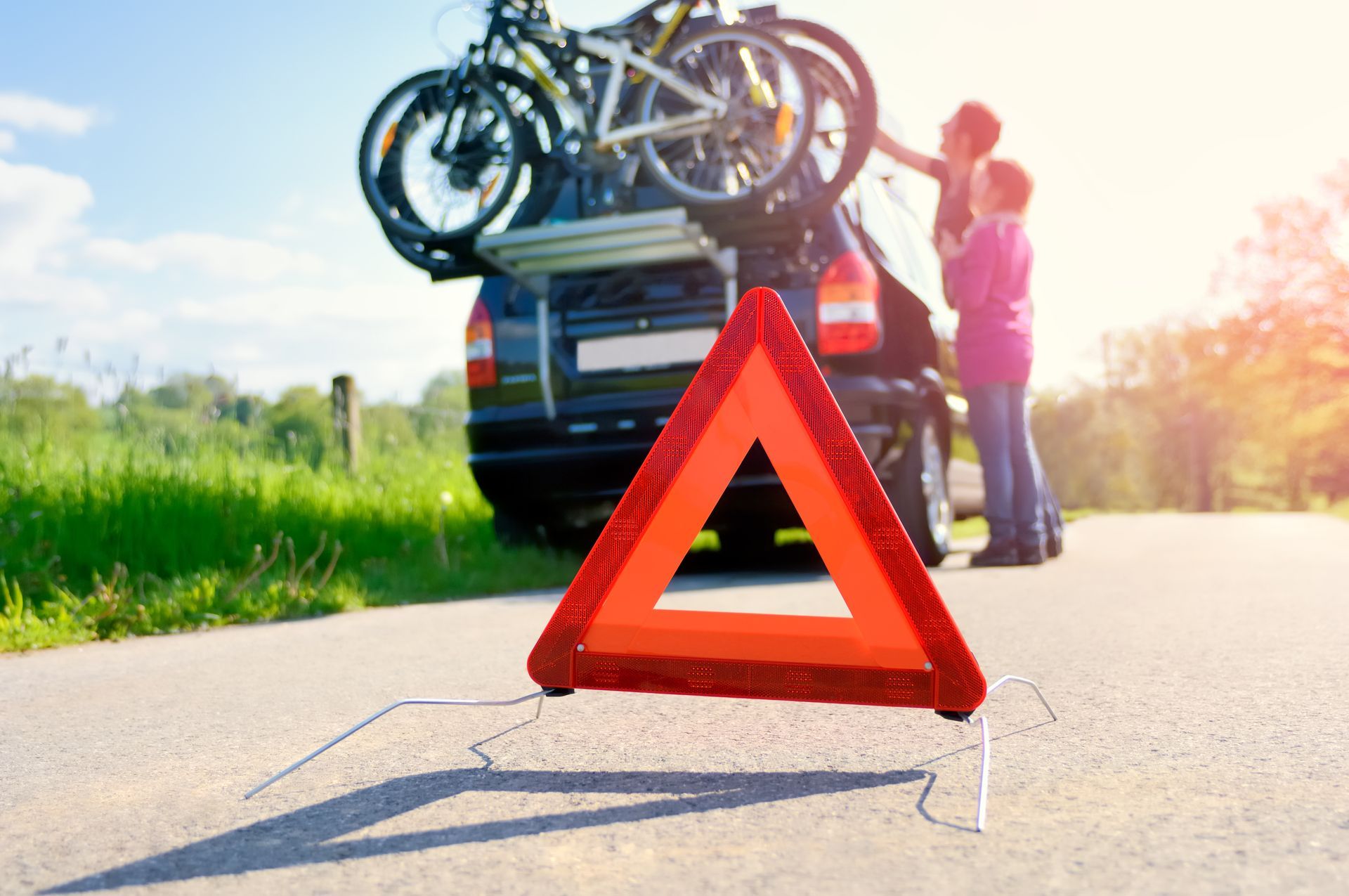 Triangle de signalisation routière au premier plan ; des personnes chargent des vélos sur une voiture en bord de route ensoleillée.