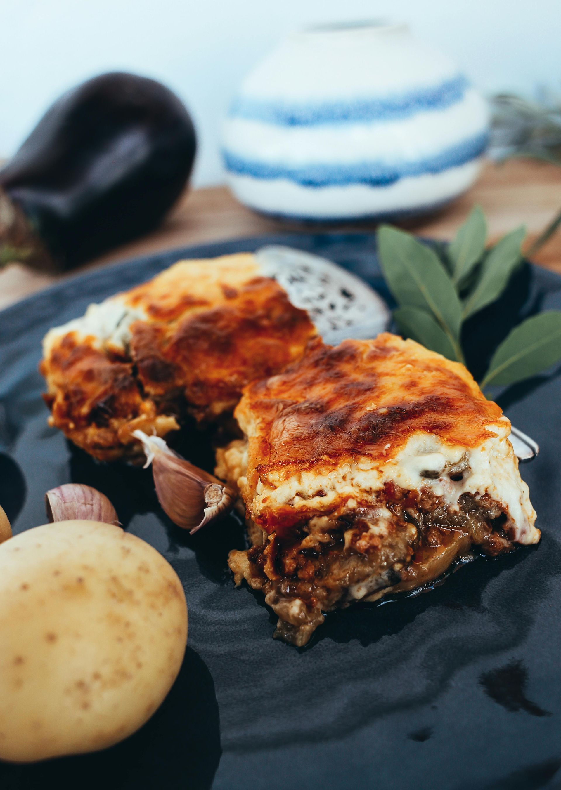 Lasaña en un plato oscuro con champiñones, patatas y hojas de albahaca, delante de un cuenco a rayas.