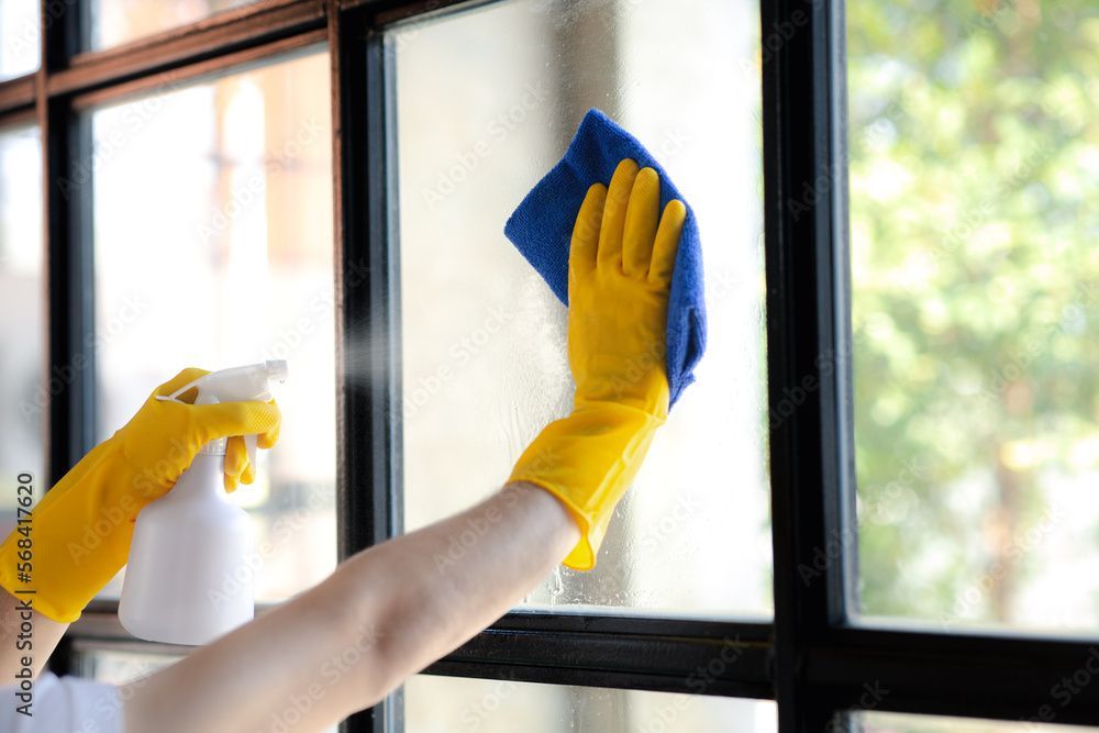 Hände in gelben Handschuhen reinigen ein Fenster mit einer Sprühflasche und einem blauen Tuch.