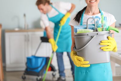 Haushälterinnen mit Reinigungsmitteln, eine wischt den Boden, die andere hält einen Putzeimer in einer hellen Küche