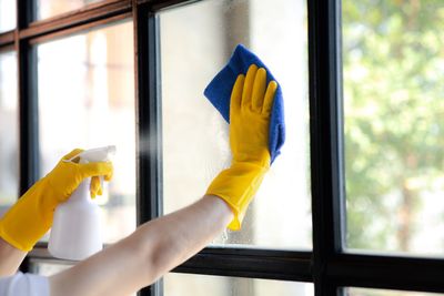 Hände in gelben Handschuhen reinigen ein Fenster mit einer Sprühflasche und einem blauen Tuch.