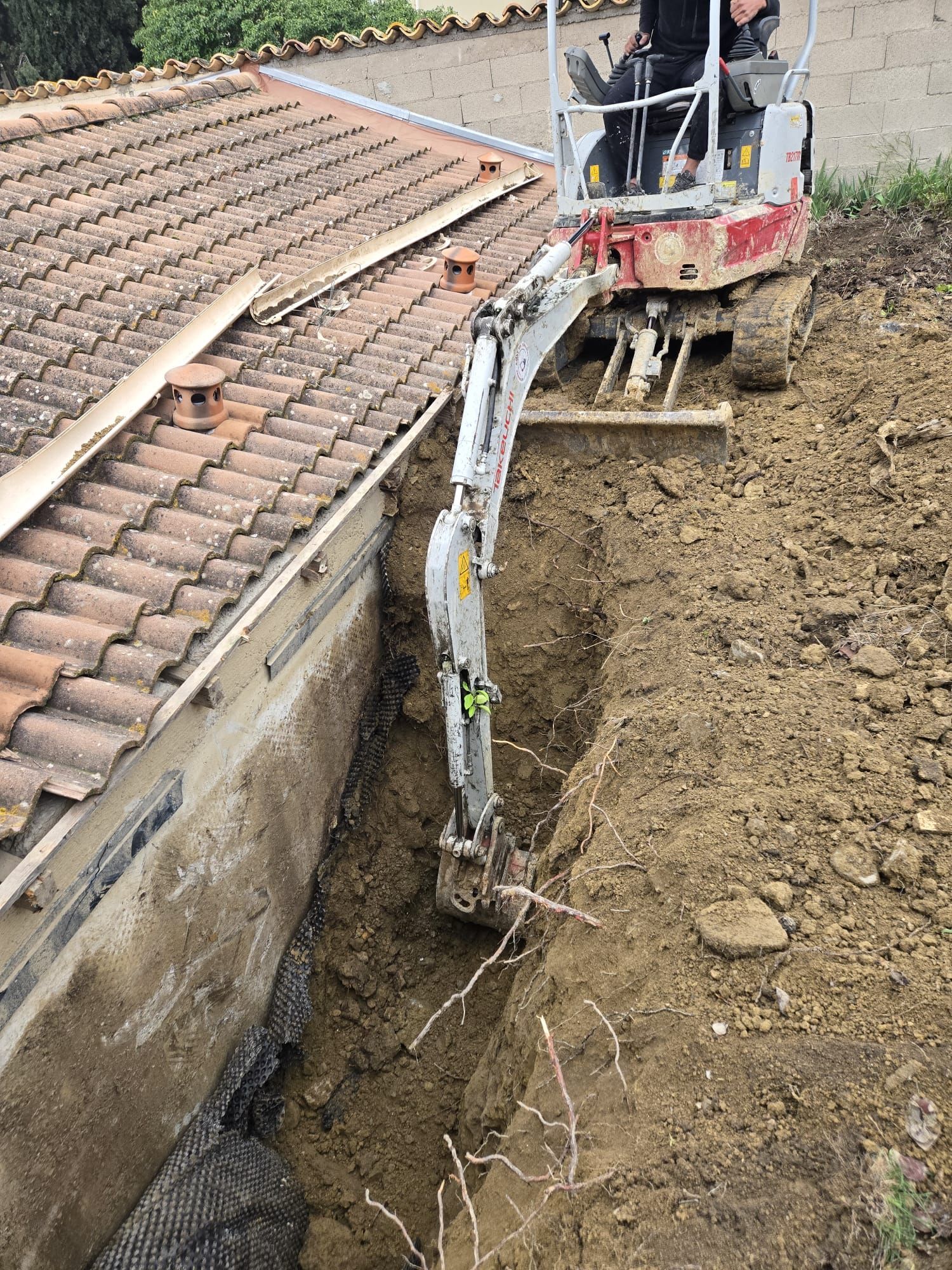 Une mini-pelle rouge creuse une tranchée juste à côté des fondations d'un bâtiment au toit de tuiles.
