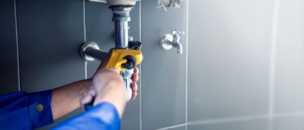 Une personne en uniforme bleu utilise une clé à pipe jaune pour serrer un tuyau métallique sous un évier contre un mur carrelé.