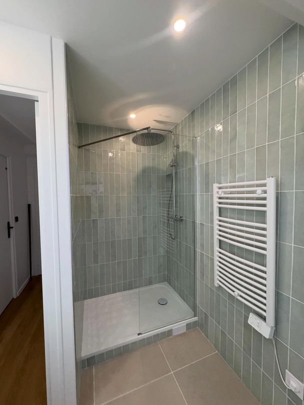 Petite salle de bains carrelée avec cabine de douche en verre, sol beige et sèche-serviettes blanc.