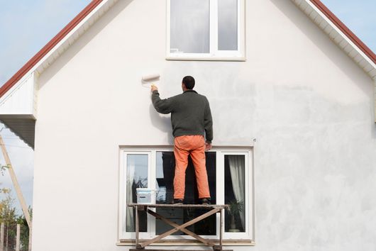 Un homme peint le mur extérieur d'une maison, debout sur un échafaudage.