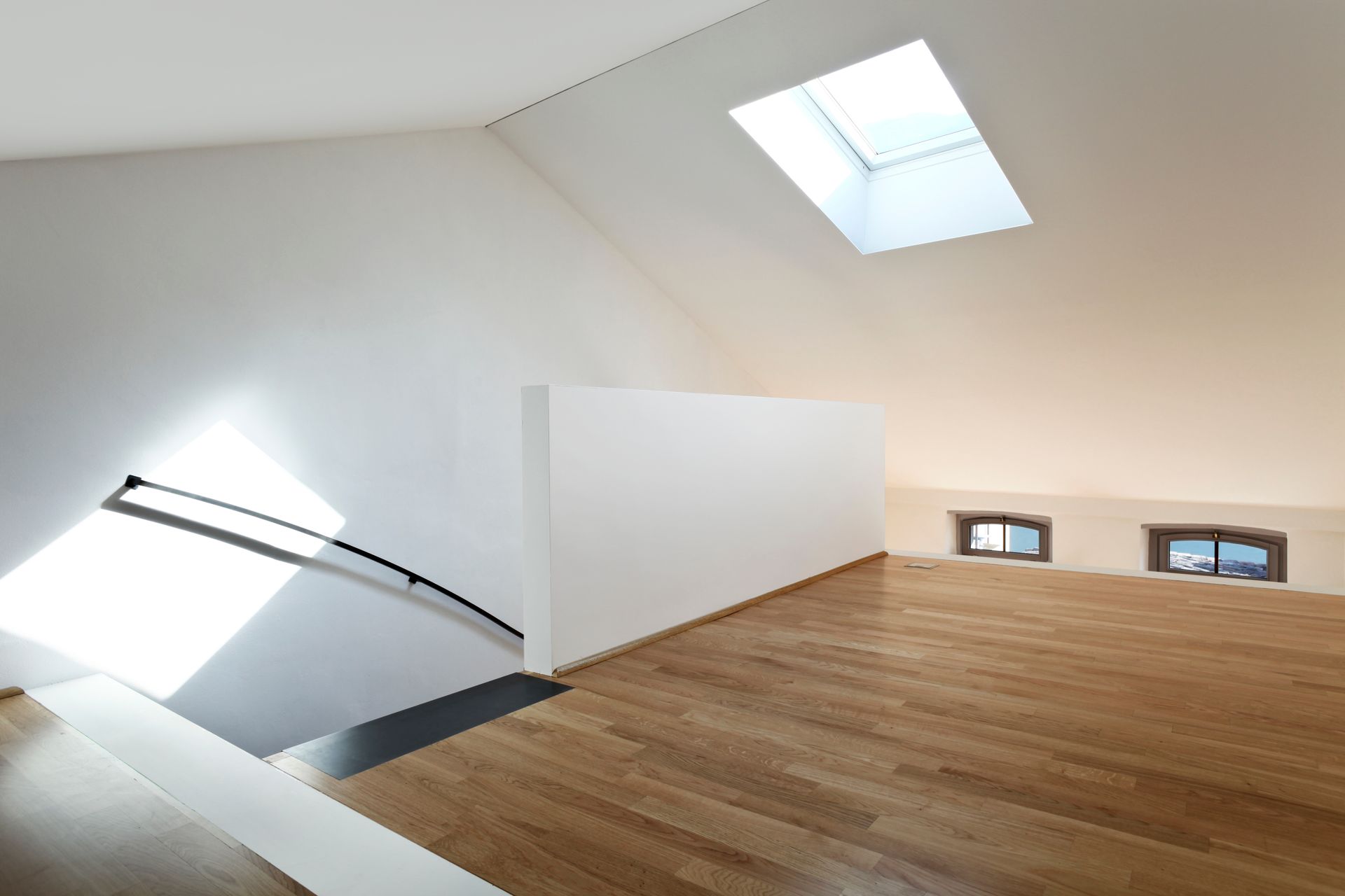 Intérieur lumineux sous les toits, avec murs blancs inclinés, puits de lumière, parquet et rampe d'escalier.