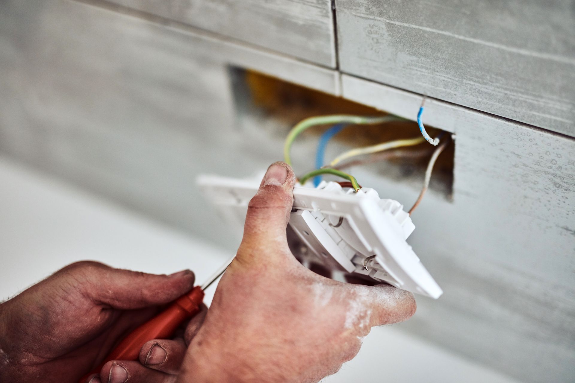 Des mains installent une prise électrique blanche dans un mur, les fils sont apparents.