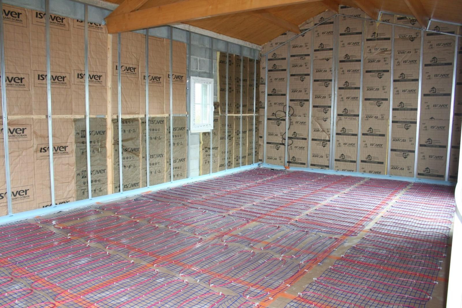 Intérieur d'une pièce avec montants muraux apparents, isolation et tubes de chauffage radiant rouges au sol.