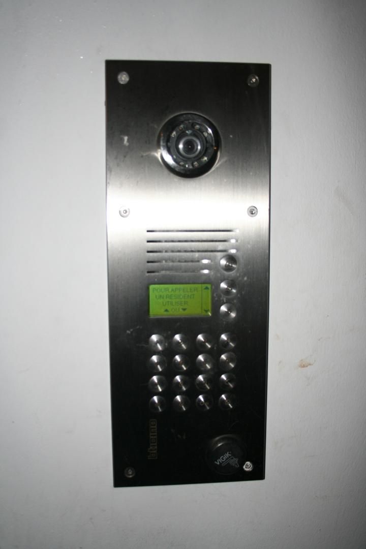 Interphone argenté avec caméra, clavier et écran LCD, monté sur un mur blanc.