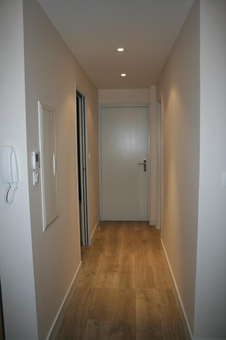 Couloir étroit aux murs beiges, au sol imitation bois et avec une porte fermée au fond.