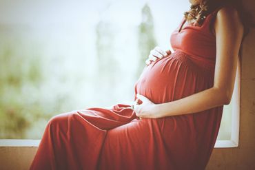 Eine schwangere Frau in einem roten Kleid sitzt auf einem Fensterbrett und hält ihren Bauch.
