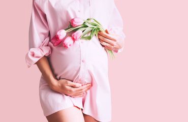 Eine schwangere Frau in einem rosa Hemd hält einen Strauß rosa Blumen in der Hand.
