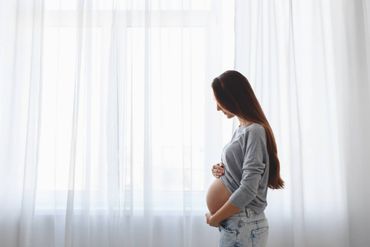 Eine schwangere Frau steht vor einem Fenster und hält ihren Bauch.