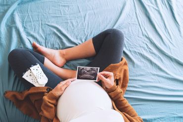 Eine schwangere Frau sitzt auf einem Bett und hält ein Ultraschallbild ihres Babys in der Hand.