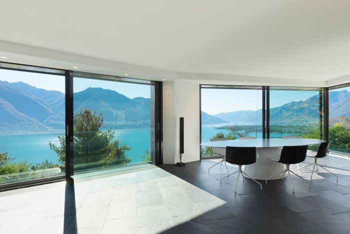 Habitación luminosa con grandes ventanales con vistas a un lago y a las montañas; mesa redonda y sillas.