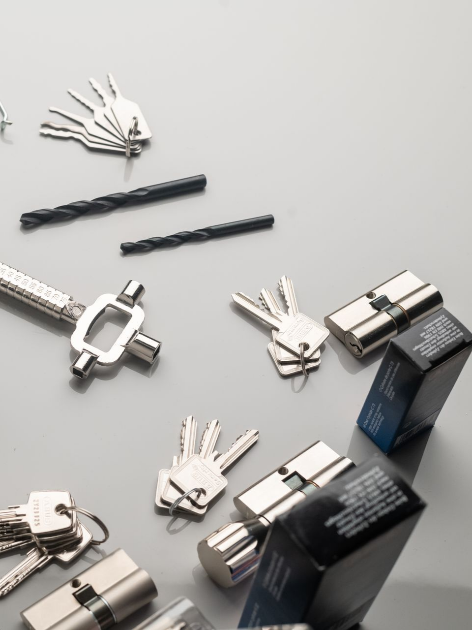 Des clés, des cylindres de serrure, des forets et un outil sur une surface grise, correspondant à des outils de serrurier.