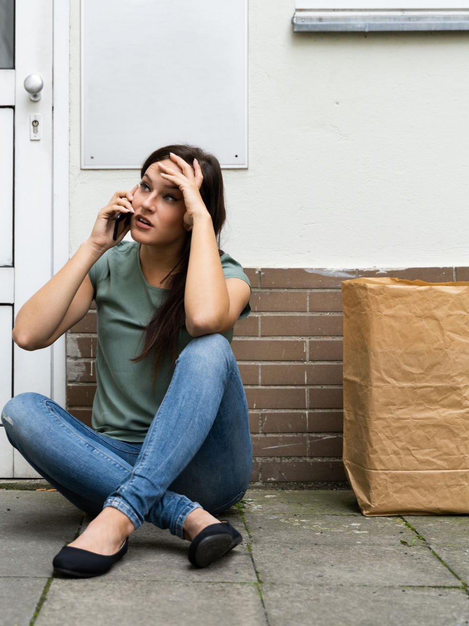Femme assise par terre, parlant au téléphone, l'air stressé, avec un sac en papier à proximité.