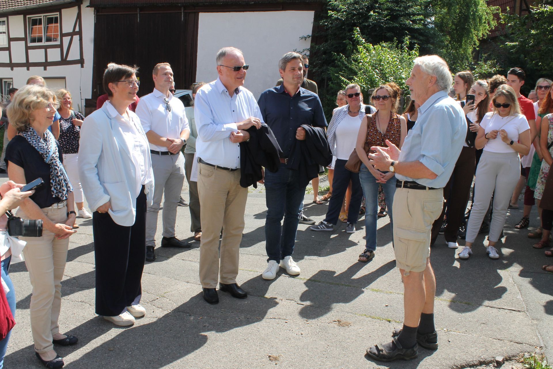 Eine Gruppe von Menschen steht draußen und hört einem Mann zu, der spricht. Einige tragen Sonnenbrillen und helle Kleidung.