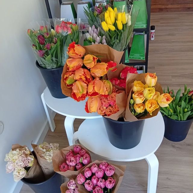 Des bouquets de fleurs colorées dans des seaux et des vases, disposés sur une table blanche dans un magasin ou un marché.