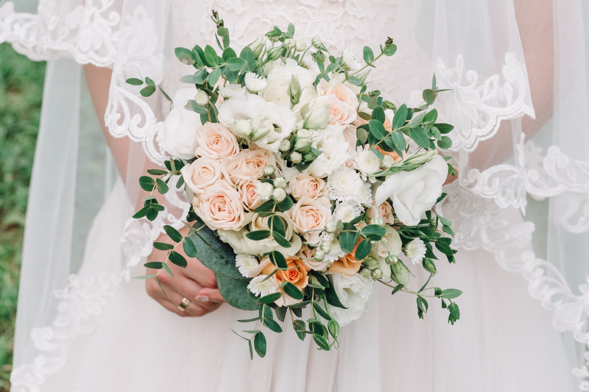 La mariée tient un bouquet crème et pêche avec de la verdure devant sa robe de mariée en dentelle blanche.