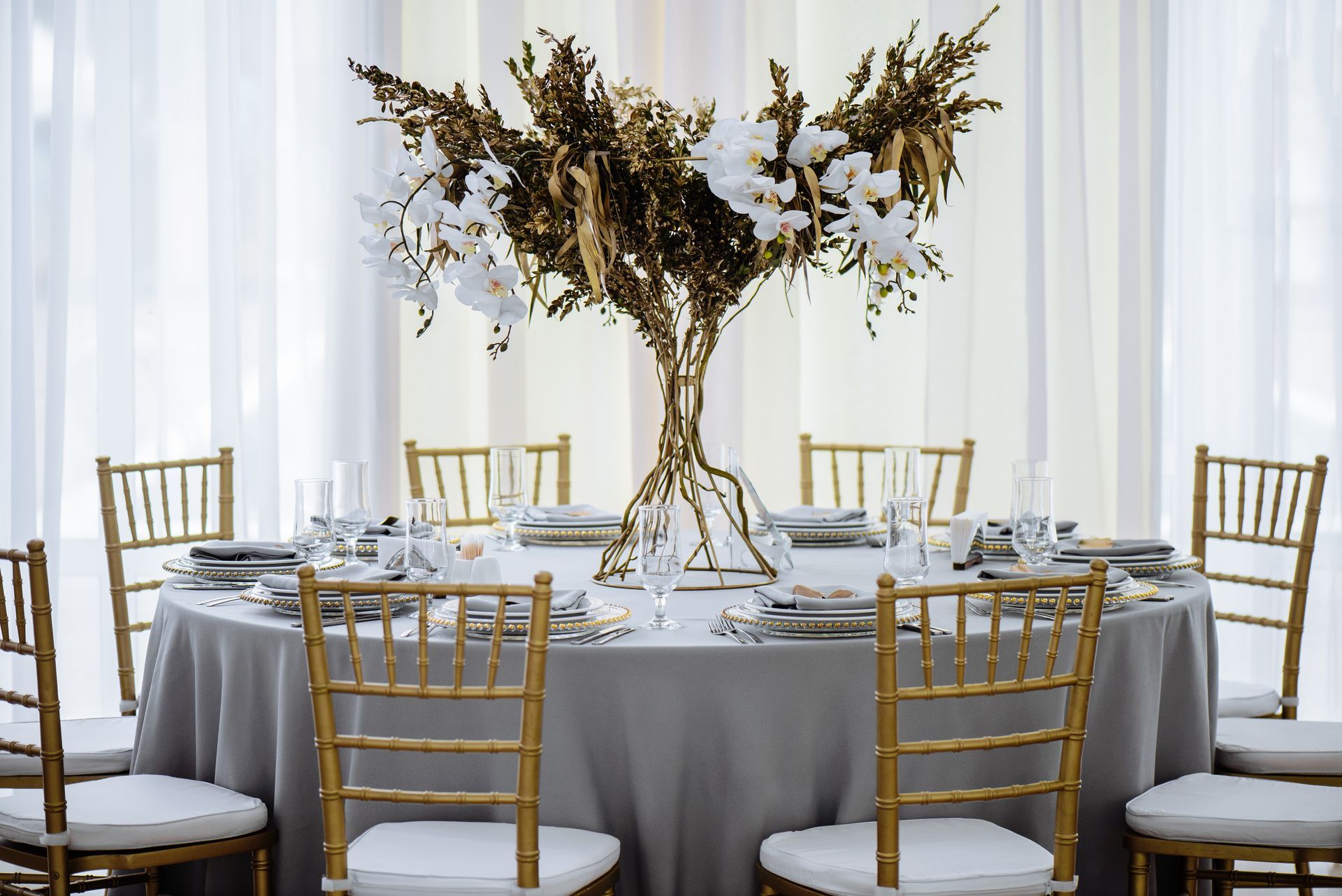 Élégante table de banquet ronde avec nappes blanches, chaises dorées et un grand centre de table floral dans une salle lumineuse