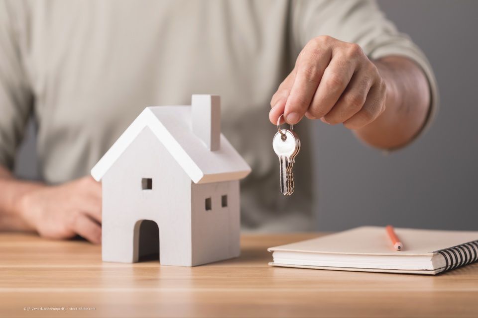 Person hält Schlüssel ins Bild, daneben ein Modellhaus