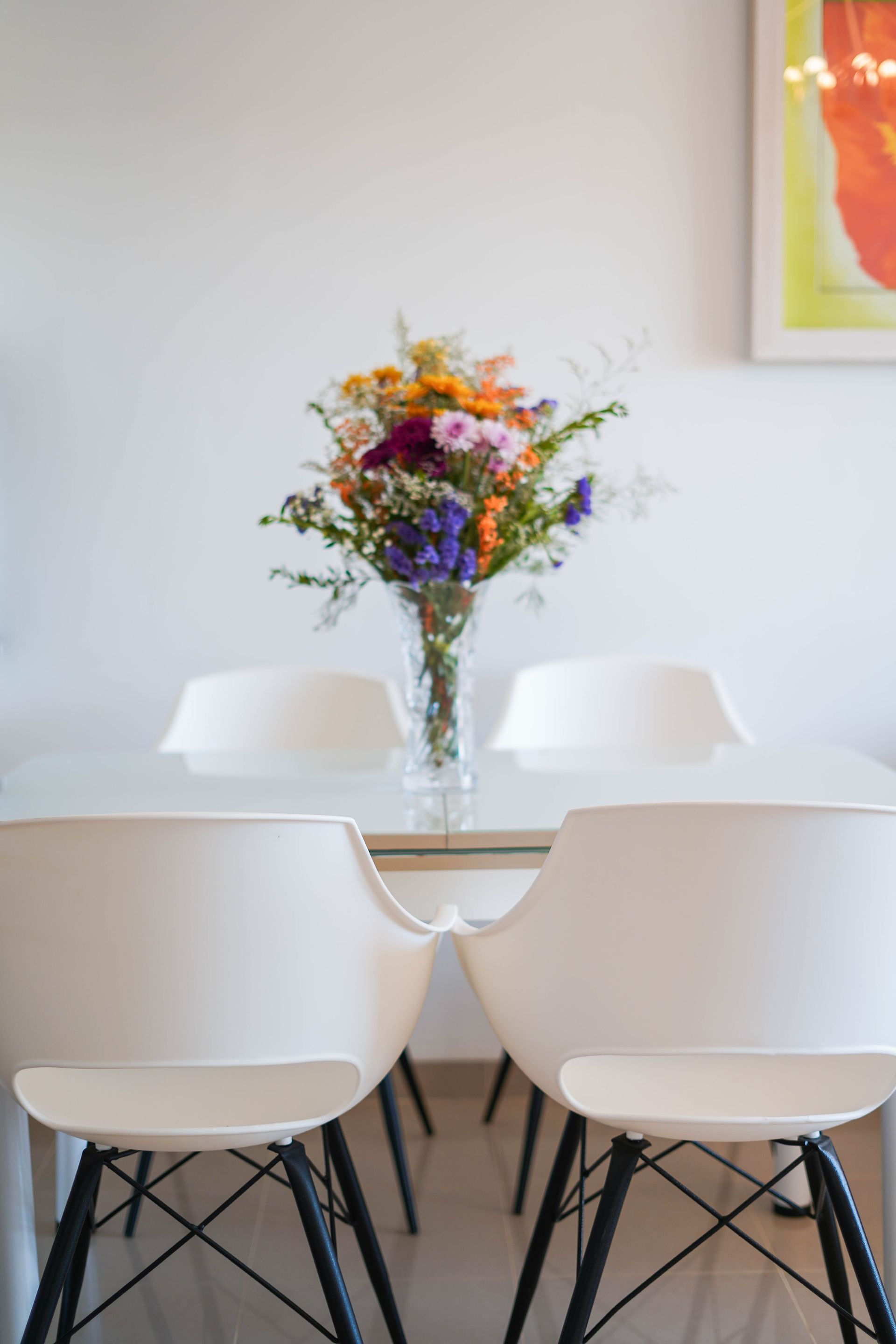 White dining table with a colorful flower bouquet in a glass vase; white chairs with black legs.