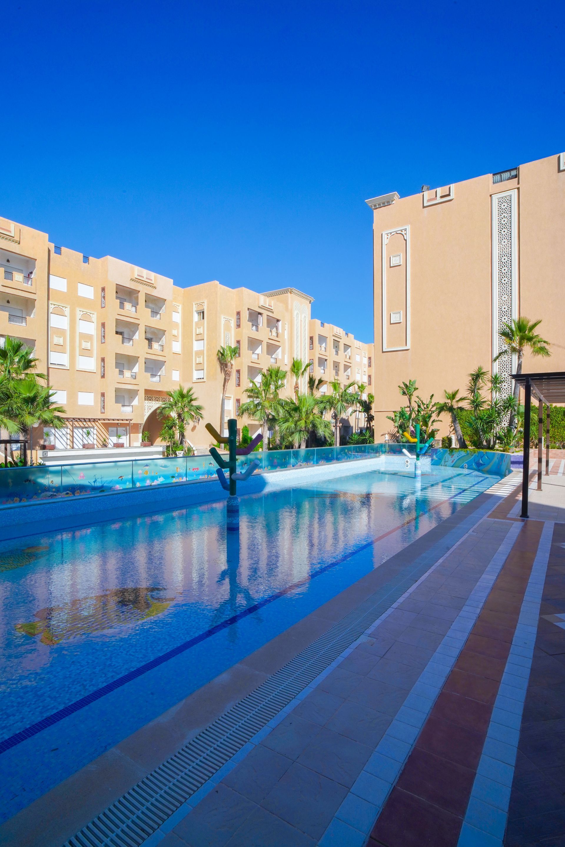 Swimming pool in front of tan buildings under a bright blue sky. Palm trees and patio visible.