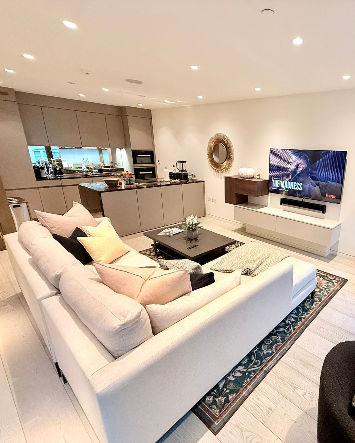 Modern living room with kitchen. Light-colored sectional sofa, TV, rug. Beige kitchen cabinets, round mirror.