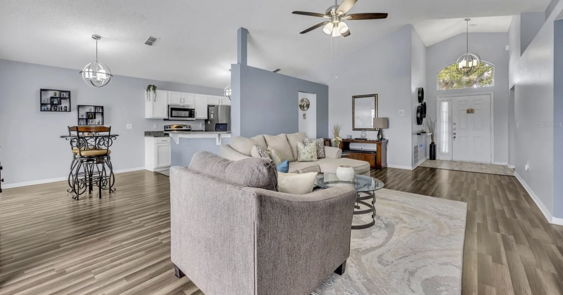 Spacious living room with gray walls, sofa, and hardwood floors. Kitchen visible in the background.
