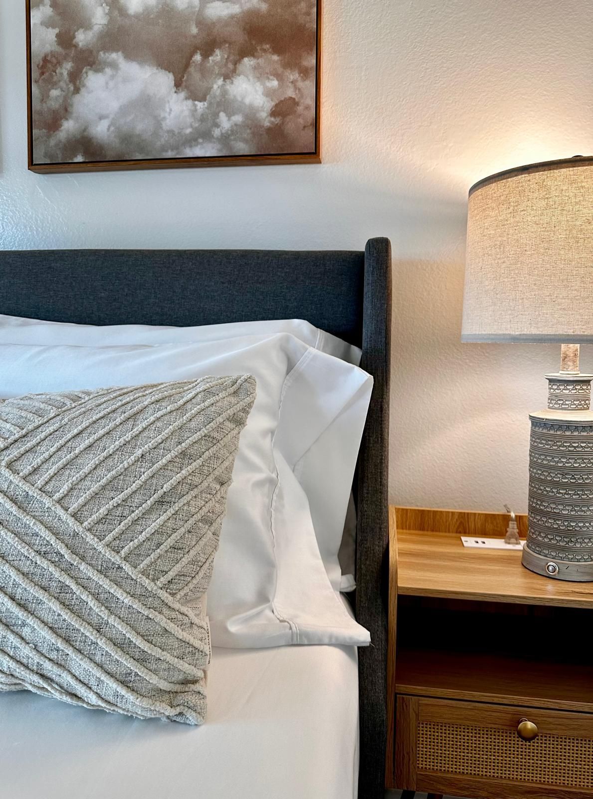 Bed with gray headboard, white sheets, decorative pillow, wooden nightstand, and lit lamp.