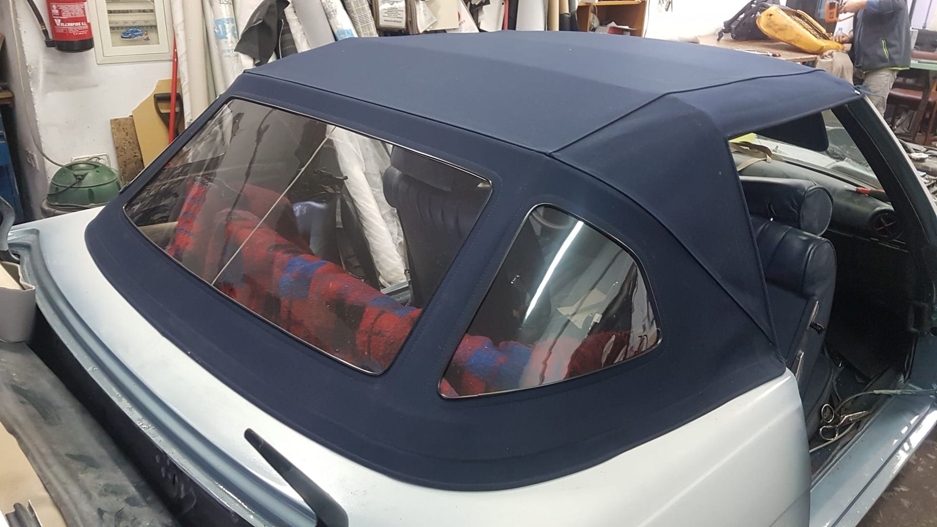 Coche descapotable con capota de lona azul oscuro en un garaje, donde se aprecian la luneta trasera y la carrocería plateada.