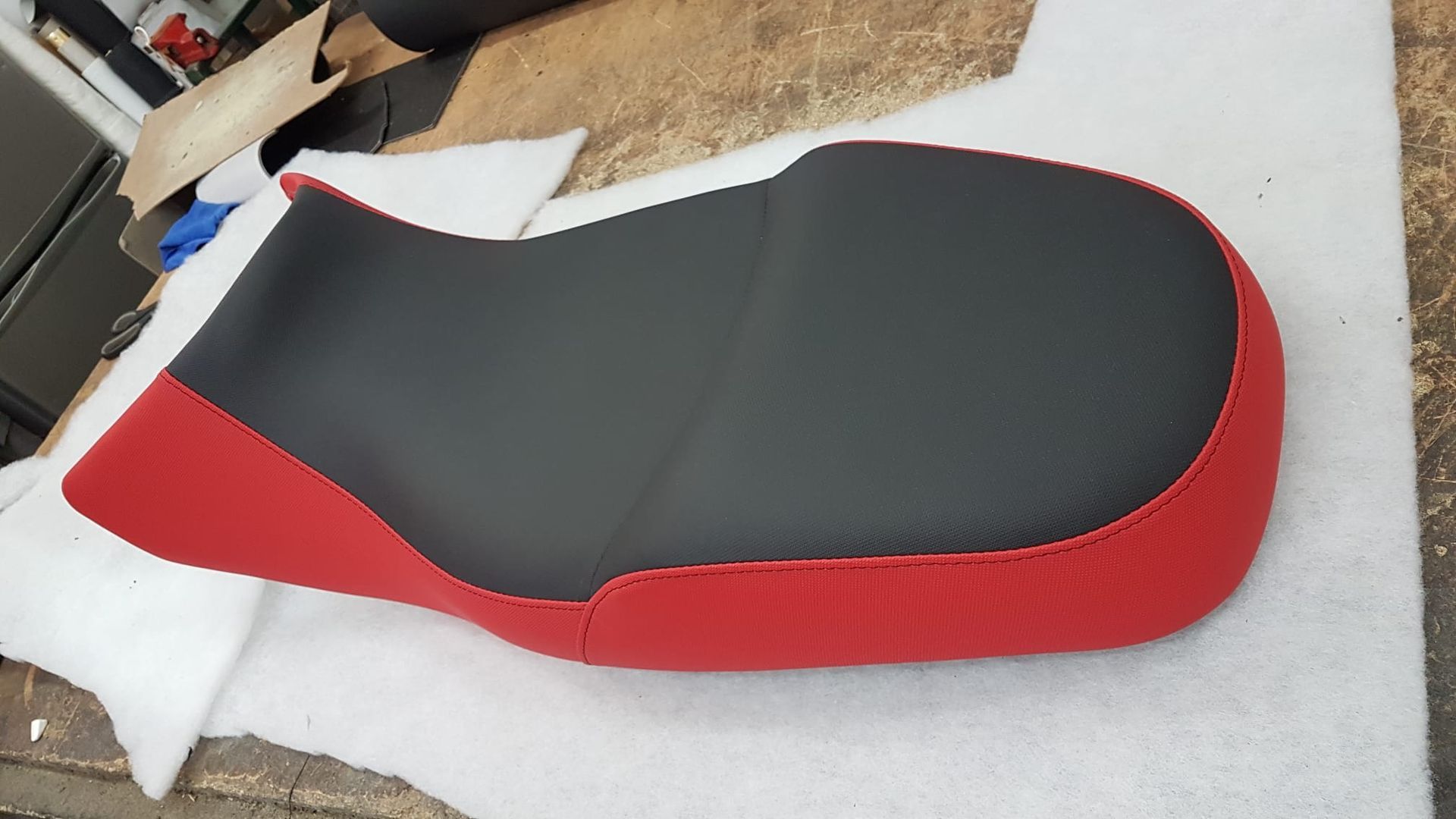 Asiento de motocicleta rojo y negro sobre una superficie blanca con objetos de taller cerca.